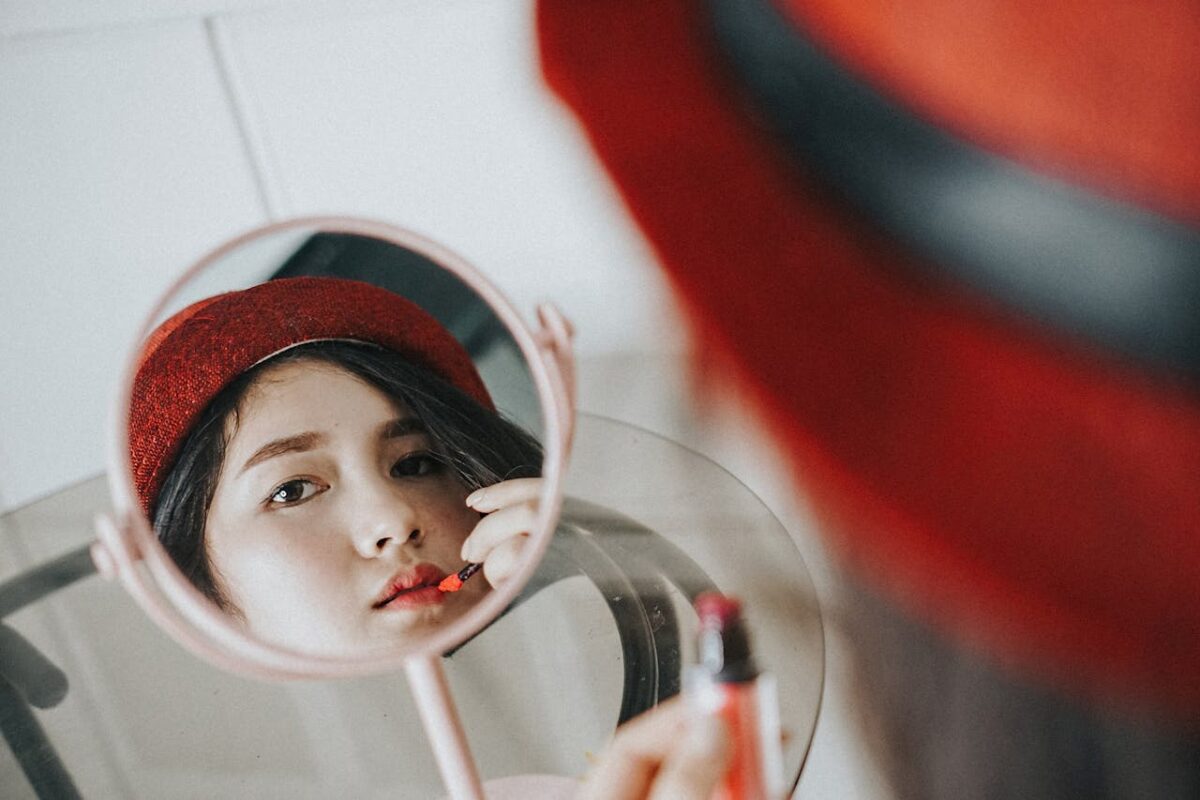 Close-up of a woman applying lipstick in front of a mirror, wearing a red hat.