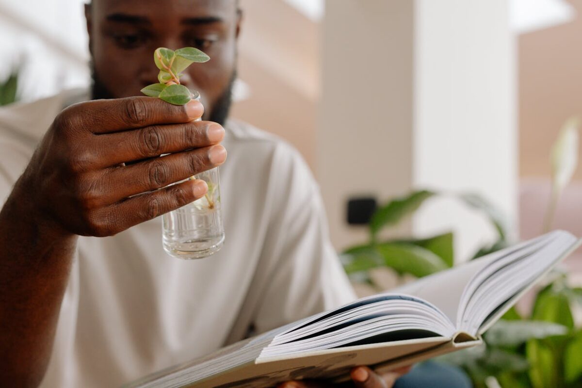 A man holds a small plant while reading a book indoors, emphasizing learning and nature.